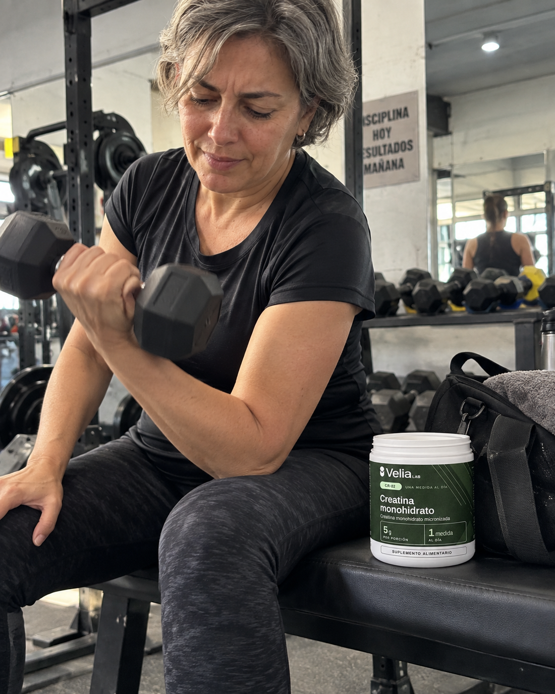 Mujer de unos 55 años entrenando con mancuerna en el gimnasio, con el pote CR-02 en la banca