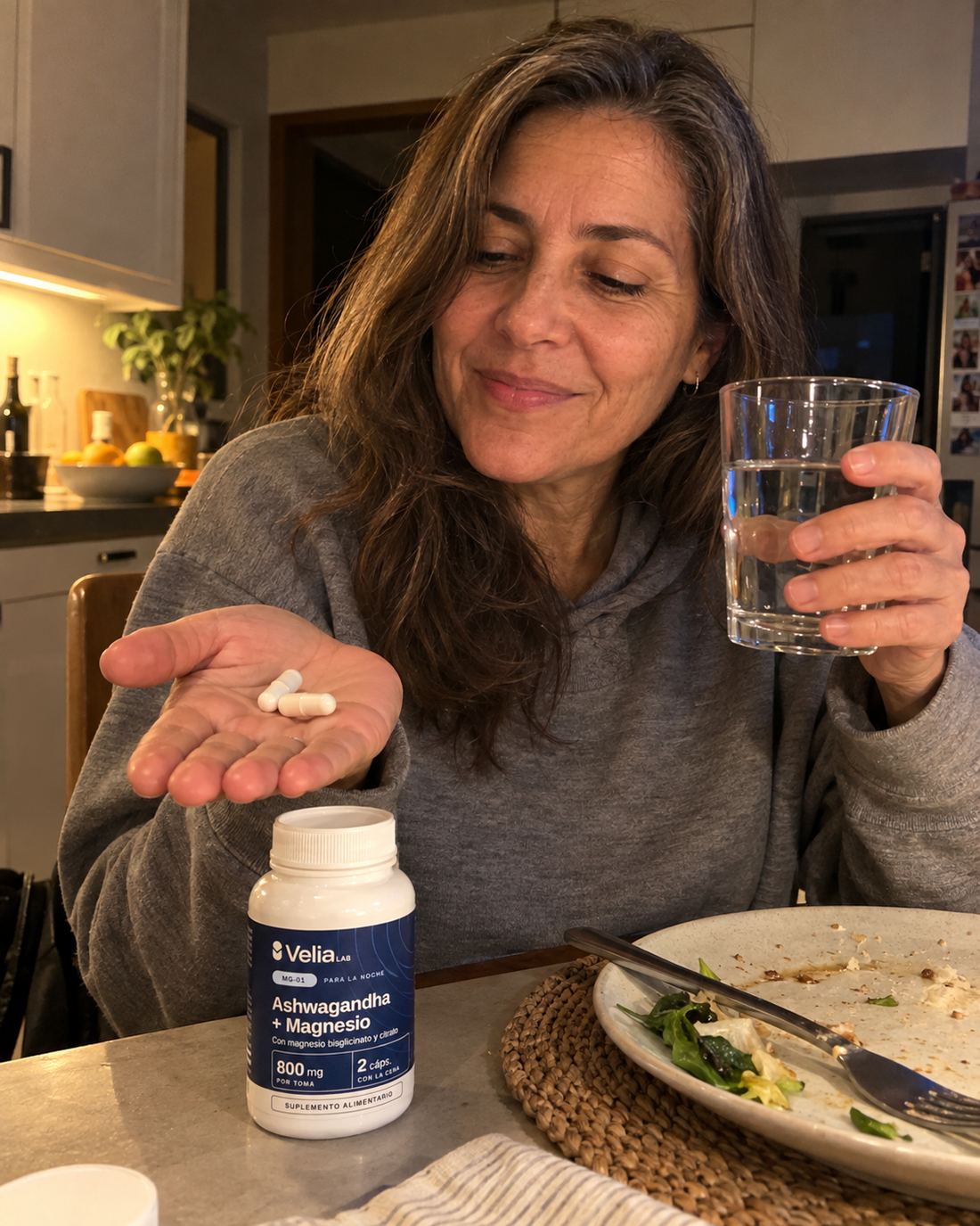 Mujer en la cocina con dos cápsulas MG-01 en la mano y un vaso de agua, después de cenar