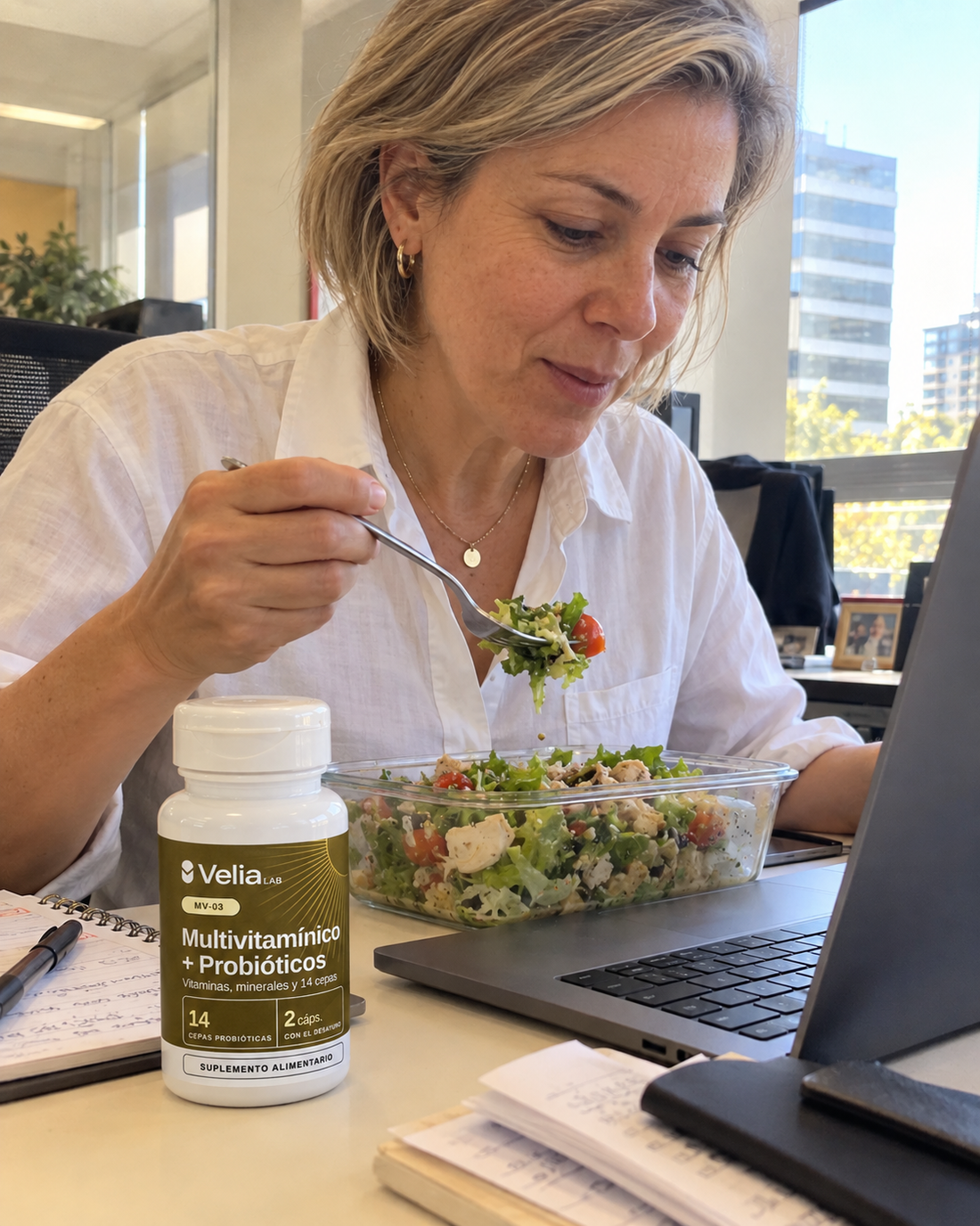 Mujer almorzando una ensalada frente al computador, con el frasco MV-03 en el escritorio